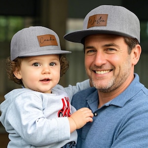 Op de afbeelding: Een vader en zoon dragen bijpassende grijze baseballpetten met gepersonaliseerde leren patches. De pet van de vader heeft de tekst "Weston" en die van de zoon "Camden".