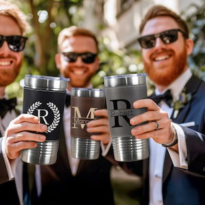 Puede incluir: Cuatro vasos de acero inoxidable personalizados con envolturas de imitaci&oacute;n de cuero. Los vasos est&aacute;n en manos de cuatro personas. Los vasos est&aacute;n grabados con los nombres "Robert", "Marc" y "R".