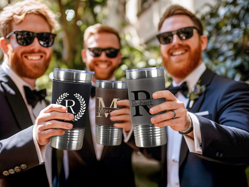 Puede incluir: Cuatro vasos de acero inoxidable personalizados con envolturas de imitaci&oacute;n de cuero. Los vasos est&aacute;n en manos de cuatro personas. Los vasos est&aacute;n grabados con los nombres "Robert", "Marc" y "R".