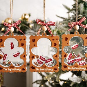 May include: Three gingerbread man-shaped Christmas ornaments. Each ornament has a clear center with small, personalized name tags and gingerbread cookies. The ornaments are decorated with red and white accents and are hanging from twine with red and white striped bows.