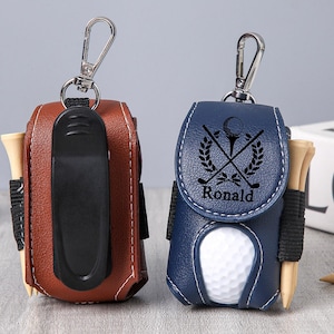 May include: Two leather golf ball pouches, one brown and one navy blue, each with a silver clip. The navy pouch has a golf-themed design and the name "Ronald". Both pouches hold golf tees.