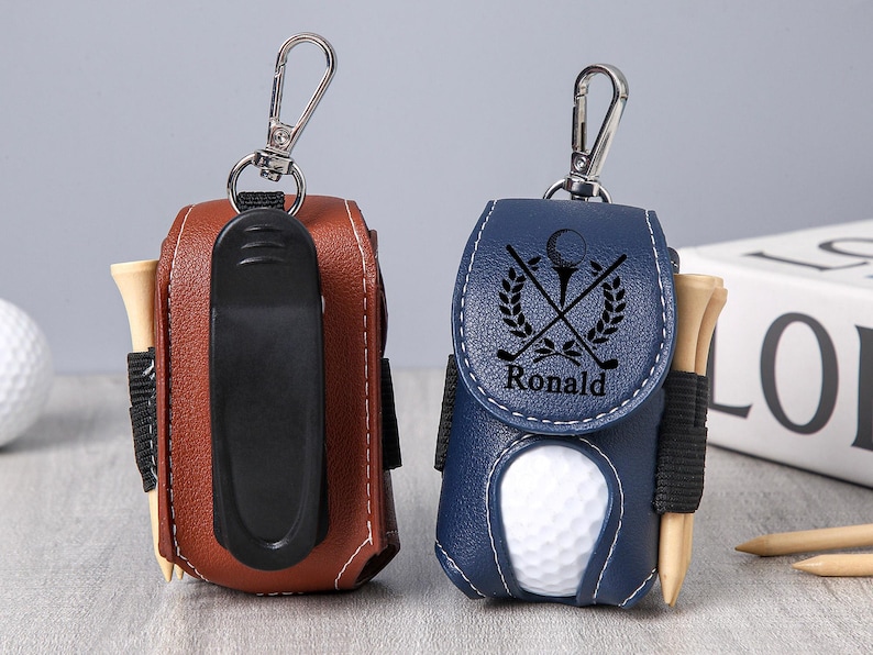 May include: Two leather golf ball pouches, one brown and one navy blue, each with a silver clip. The navy pouch has a golf-themed design and the name "Ronald". Both pouches hold golf tees.