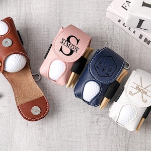 May include: A collection of personalized golf ball holders in brown, pink, navy blue, and white. Each holder contains a golf ball and tees. The pink holder has the name "SIMON" and the white holder has the name "Barton".