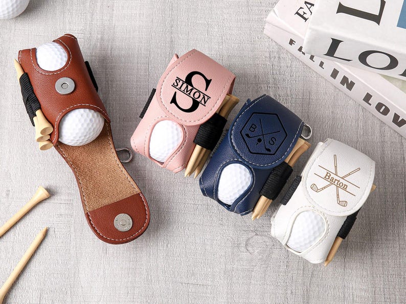 May include: A collection of personalized golf ball holders in brown, pink, navy blue, and white. Each holder contains a golf ball and tees. The pink holder has the name "SIMON" and the white holder has the name "Barton".