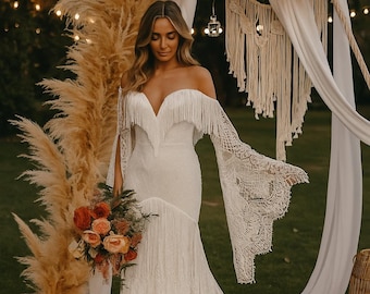 May include: An off-the-shoulder white lace wedding dress featuring a deep V-neckline, long flared bell sleeves with intricate crochet detailing, and a bodice adorned with fringe trim that extends across the chest and shoulders.