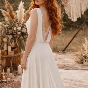 K&ouml;nnte beinhalten: Ein wei&szlig;es Hochzeitskleid mit tiefem R&uuml;cken und zarten Spitzendetails. Das &auml;rmellose Kleid hat einen flie&szlig;enden Rock und einen Knopfverschluss hinten. Das Bild ist im Freien mit einem nat&uuml;rlichen, b&ouml;hmischen Thema aufgenommen, mit einer Frau mit langen roten Haaren.