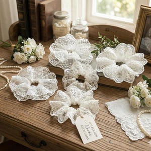 May include: A collection of white lace scrunchies with a delicate, ruffled design. The scrunchies are arranged on a wooden surface with a tag that reads "Colección Novia Reciclada". Other items include a bouquet of white flowers and a pearl necklace.