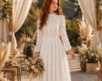 May include: A long-sleeved white lace dress featuring a high neckline, a fitted bodice, and a flowing skirt. The intricate floral lace pattern covers the entire garment, which is paired with a bouquet of mixed cream and beige flowers.