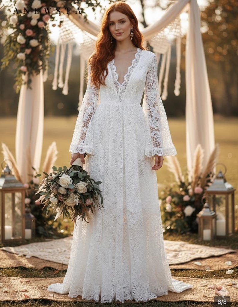 May include: A white lace wedding dress with a plunging neckline and long, flared sleeves. The dress features intricate floral lace detailing and a flowing skirt. The bride is holding a bouquet of white and green flowers. The setting is outdoors with a decorative arch.