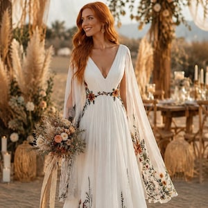 Puede incluir: Un vestido blanco largo hasta el suelo con un escote en V profundo, mangas tipo capa fluidas y un intrincado bordado floral en tonos naranja, marrón y verde a lo largo de la cintura, que cae por la falda y las mangas, de tela ligera.