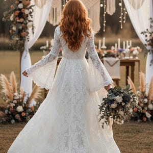 May include: A white lace wedding dress with long, flared sleeves and a detailed hem. The bride is holding a bouquet of flowers. The background includes floral arrangements and decorative macrame.