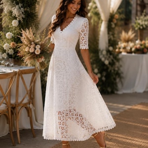 May include: A white lace midi dress with a V-neck and short sleeves. The dress features a scalloped hemline and a floral lace pattern. The person is wearing gold high heels. The background includes floral arrangements and a table setting.