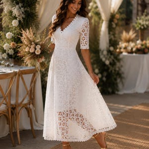May include: A white lace midi dress with a V-neck and short sleeves. The dress features a scalloped hemline and a floral lace pattern. The person is wearing gold high heels. The background includes floral arrangements and a table setting.