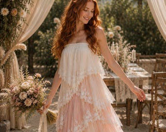 May include: A strapless tiered dress featuring layers of cream and soft pink tulle with white lace trim along the edges. The dress has a flowing silhouette and is paired with a bouquet of dried flowers in shades of cream and dusty rose.