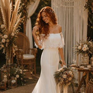 Peut inclure: Une robe de mariée blanche à épaules dénudées avec une superposition de dentelle et une silhouette sirène. La mariée tient un bouquet de fleurs. L'arrière-plan comprend une toile de fond en macramé, des compositions florales et une chaise en osier.
