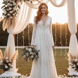 May include: A white lace wedding dress with long sleeves and a deep V-neck. The bride is holding a bouquet of white and green flowers. The ceremony backdrop includes a wooden arch, white draping fabric, and floral arrangements.
