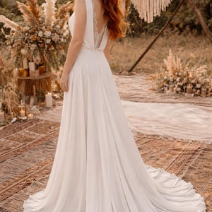 K&ouml;nnte beinhalten: Ein wei&szlig;es Hochzeitskleid mit flie&szlig;endem Rock und tiefem R&uuml;cken mit Spitzendetails. Das Kleid hat ein schlichtes, elegantes Design. Die Braut steht im Freien in einer nat&uuml;rlichen Umgebung, mit einer Kulisse aus Blumen und Gr&uuml;n.