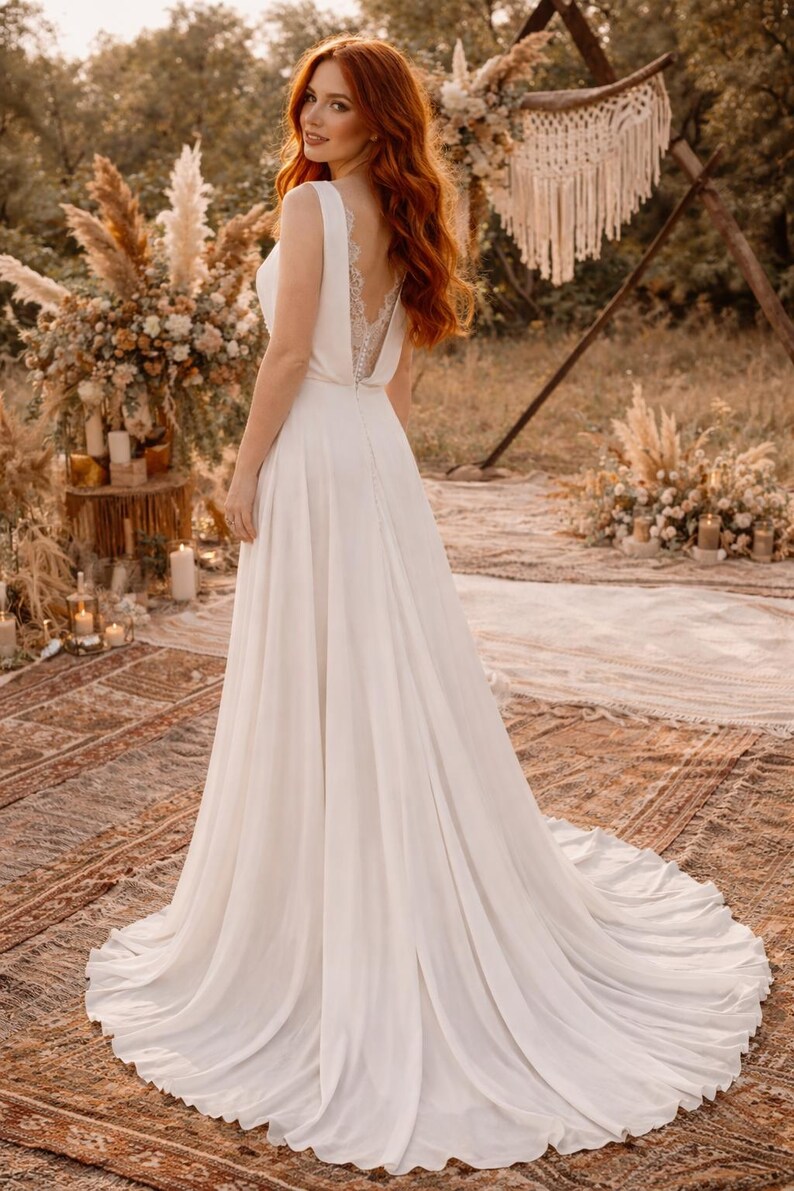 K&ouml;nnte beinhalten: Ein wei&szlig;es Hochzeitskleid mit flie&szlig;endem Rock und tiefem R&uuml;cken mit Spitzendetails. Das Kleid hat ein schlichtes, elegantes Design. Die Braut steht im Freien in einer nat&uuml;rlichen Umgebung, mit einer Kulisse aus Blumen und Gr&uuml;n.