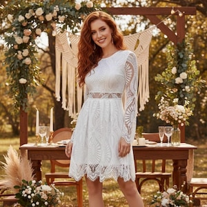 May include: A white lace dress with long sleeves and a sheer waist. The dress has a scalloped hem and is worn outdoors. The background includes a wooden table, flowers, and a decorative arch, suggesting a wedding or special event.