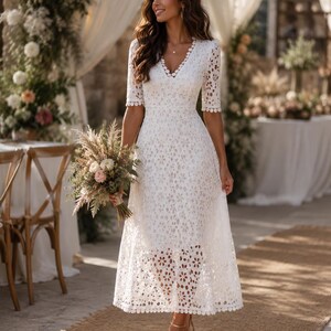 May include: A white, knee-length dress with a floral lace pattern. The dress features short sleeves and a V-neckline. The person is holding a bouquet of flowers and wearing gold-colored heels. The background includes floral arrangements and a table.