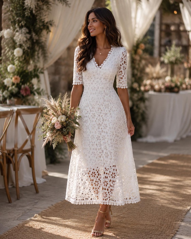 May include: A white, knee-length dress with a floral lace pattern. The dress features short sleeves and a V-neckline. The person is holding a bouquet of flowers and wearing gold-colored heels. The background includes floral arrangements and a table.
