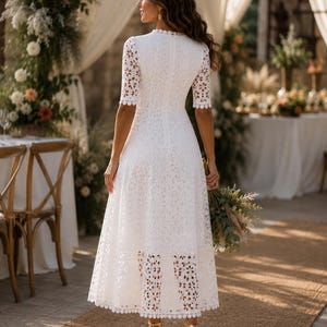 May include: A white lace midi dress with short sleeves and a scalloped neckline. The dress features a floral pattern and is paired with gold heels. The background includes floral arrangements and a table setting, suggesting a wedding or special event.
