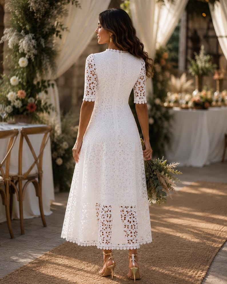 May include: A white lace midi dress with short sleeves and a scalloped neckline. The dress features a floral pattern and is paired with gold heels. The background includes floral arrangements and a table setting, suggesting a wedding or special event.