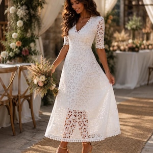 May include: A white lace dress with a V-neck and short sleeves. The dress has a floral pattern and a scalloped hem. The person is wearing gold high heels and holding a bouquet of flowers. The background includes a table and floral decorations.