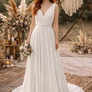 K&ouml;nnte beinhalten: Ein wei&szlig;es Brautkleid mit V-Ausschnitt und flie&szlig;endem Rock. Das &auml;rmellose Kleid hat ein eng anliegendes Oberteil. Die Braut h&auml;lt einen Blumenstrau&szlig;. Der Hintergrund zeigt Blumenarrangements und dekorative Elemente.