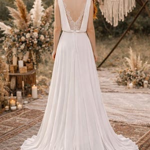 K&ouml;nnte beinhalten: Wei&szlig;es Brautkleid mit flie&szlig;endem Rock und V-Ausschnitt. Der R&uuml;cken des Kleides hat ein zartes Spitzendesign. Die Braut steht im Freien mit einem nat&uuml;rlichen Hintergrund, einschlie&szlig;lich Blumenarrangements und Kerzen.