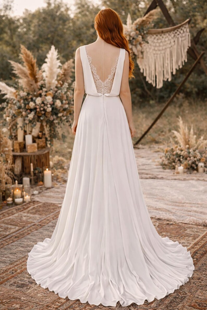 K&ouml;nnte beinhalten: Wei&szlig;es Brautkleid mit flie&szlig;endem Rock und V-Ausschnitt. Der R&uuml;cken des Kleides hat ein zartes Spitzendesign. Die Braut steht im Freien mit einem nat&uuml;rlichen Hintergrund, einschlie&szlig;lich Blumenarrangements und Kerzen.