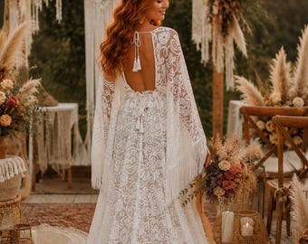 May include: A white lace wedding dress with a flowing train and open back. The dress features intricate floral patterns and fringe details on the sleeves. The bride is holding a bouquet of flowers.