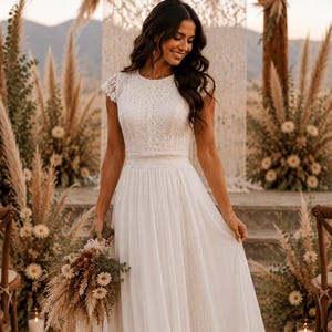 May include: A white wedding dress with a lace bodice and cap sleeves. The dress has a flowing skirt and is held by a person. The background features floral arrangements and a wooden arch, suggesting an outdoor ceremony.