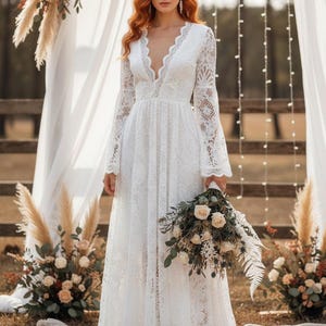 May include: A white lace wedding dress with a plunging neckline and long sleeves. The dress features a flowing A-line skirt and is held by a person. The person is holding a bouquet of white and green flowers. The dress is suitable for a wedding.