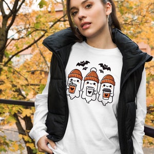 以下が含まれることがあります： ハロウィーンをテーマにしたデザインの白い長袖Tシャツ。オレンジ色の帽子をかぶり、コーヒーカップを持った3匹の漫画の幽霊が描かれています。黒いコウモリが幽霊の上を飛んでいます。シャツは黒いベストと合わせて着用されています。