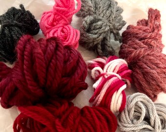 May include: Assortment of yarn in various colors, including pink, red, burgundy, and gray. The yarn is arranged in a circular pattern on a white surface.