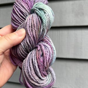 May include: A skein of hand-dyed yarn in shades of purple, lavender, and seafoam green. The yarn is held in a person's hand, showcasing the color variations and texture. The yarn appears soft and suitable for knitting or crochet projects.