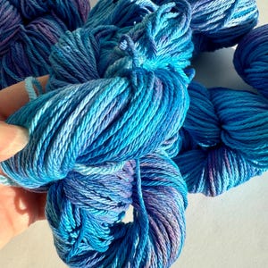 May include: Close-up of several skeins of hand-dyed yarn in shades of blue and purple. The yarn is tightly wound and has a soft, textured appearance. The colors blend together, creating a gradient effect. Ideal for knitting, crocheting, and other fiber arts projects.