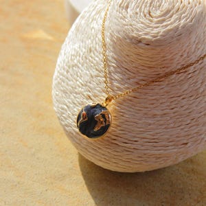 Puede incluir: Un collar dorado con un colgante redondo que representa un mapa del mundo. El colgante tiene un fondo azul oscuro con continentes dorados y detalles rojos. El collar se muestra sobre una superficie blanca texturizada.