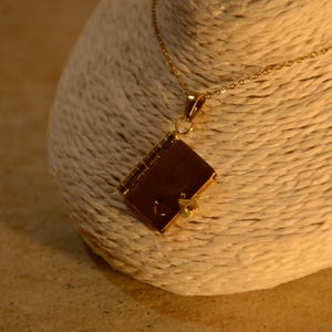 Puede incluir: Un collar con colgante dorado con un pequeño dije en forma de libro. El dije tiene un diseño detallado con un cierre y un pequeño adorno en forma de corazón. El collar se exhibe sobre una superficie texturizada de color blanquecino.