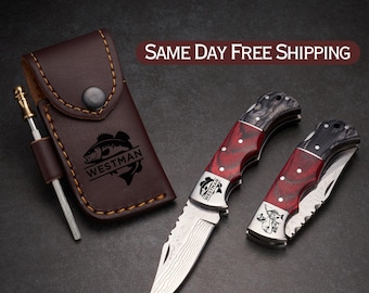 May include: A set of two folding knives with red and black handles, alongside a brown leather sheath and sharpening tool. The knives have silver blades with a textured pattern. The sheath is embossed with a fish logo and the word "WESTMAN".