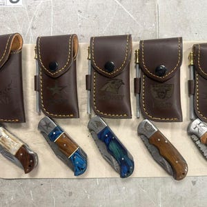 May include: Five brown leather knife sheaths with brass fire starters and matching folding pocket knives. Each sheath is embossed with names or logos including Jesse, a star, Rodney, Raiders Dad, and Kuhn. The knives feature varied handle materials.