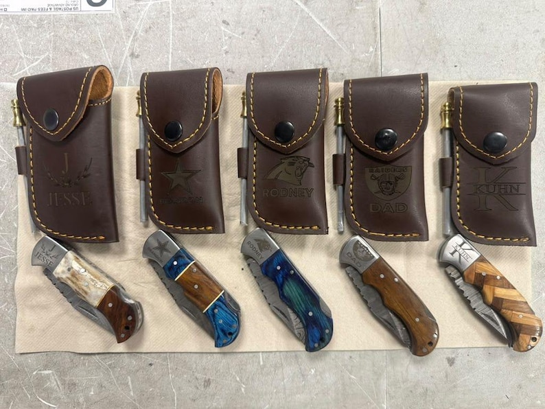 May include: Five brown leather knife sheaths with brass fire starters and matching folding pocket knives. Each sheath is embossed with names or logos including Jesse, a star, Rodney, Raiders Dad, and Kuhn. The knives feature varied handle materials.