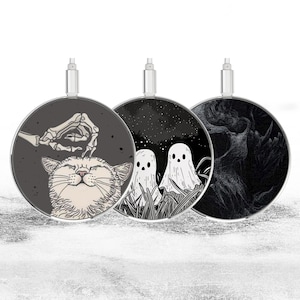 May include: Three round wireless charging pads with different black and white designs. The first pad features a cat with a skeleton hand petting it. The second pad has two ghosts in a field of grass. The third pad has a black and white abstract design.