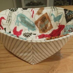 May include: A white fabric bowl with a farmhouse theme print. The bowl features a white background with a black and white checkered pattern, red and blue farm animals, and the text "Farm Sweet Farm".