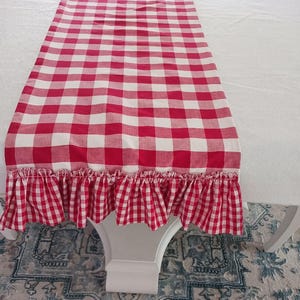May include: Red and white gingham table runner with a ruffled edge. The runner is made of cotton fabric and has a checkered pattern.