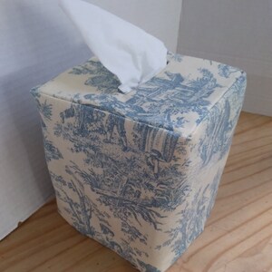 May include: A blue and white toile patterned tissue box cover. The cover is square and has a tissue box opening on the top.