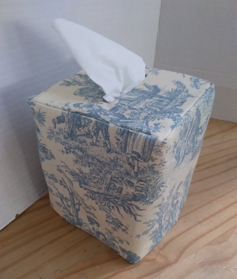 May include: A blue and white toile patterned tissue box cover. The cover is square and has a tissue box opening on the top.