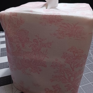 May include: A white tissue box cover with a pink floral pattern. The cover is made of fabric and has a square shape.