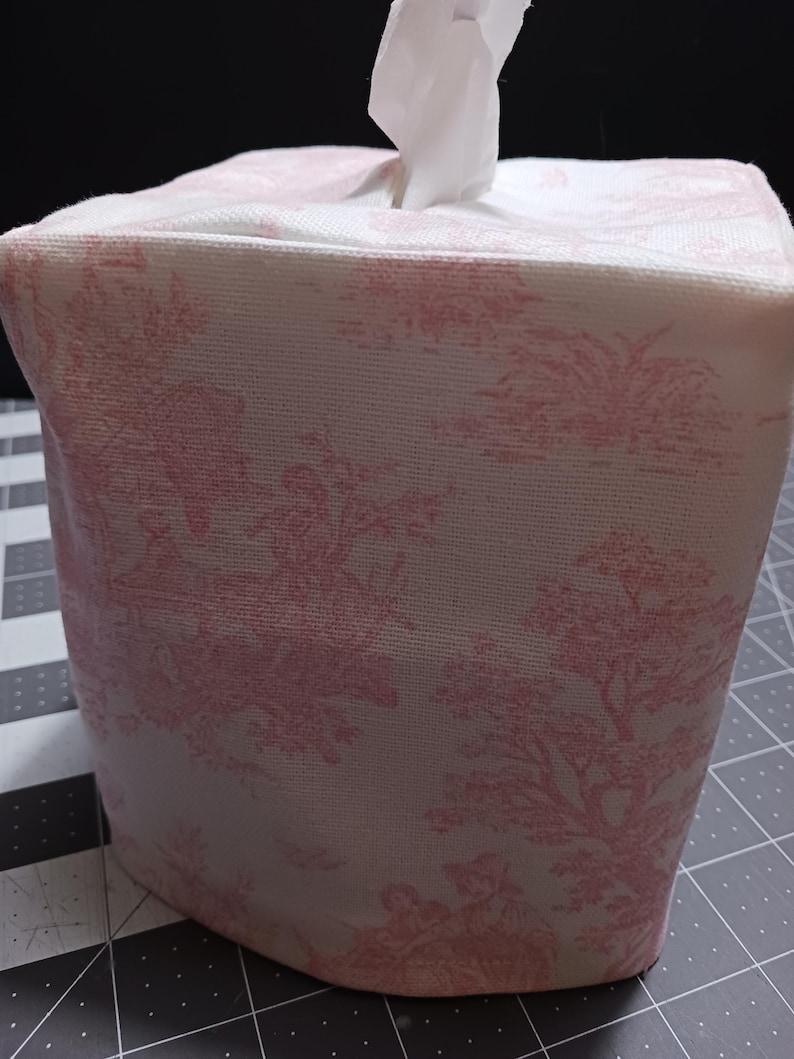 May include: A white tissue box cover with a pink floral pattern. The cover is made of fabric and has a square shape.
